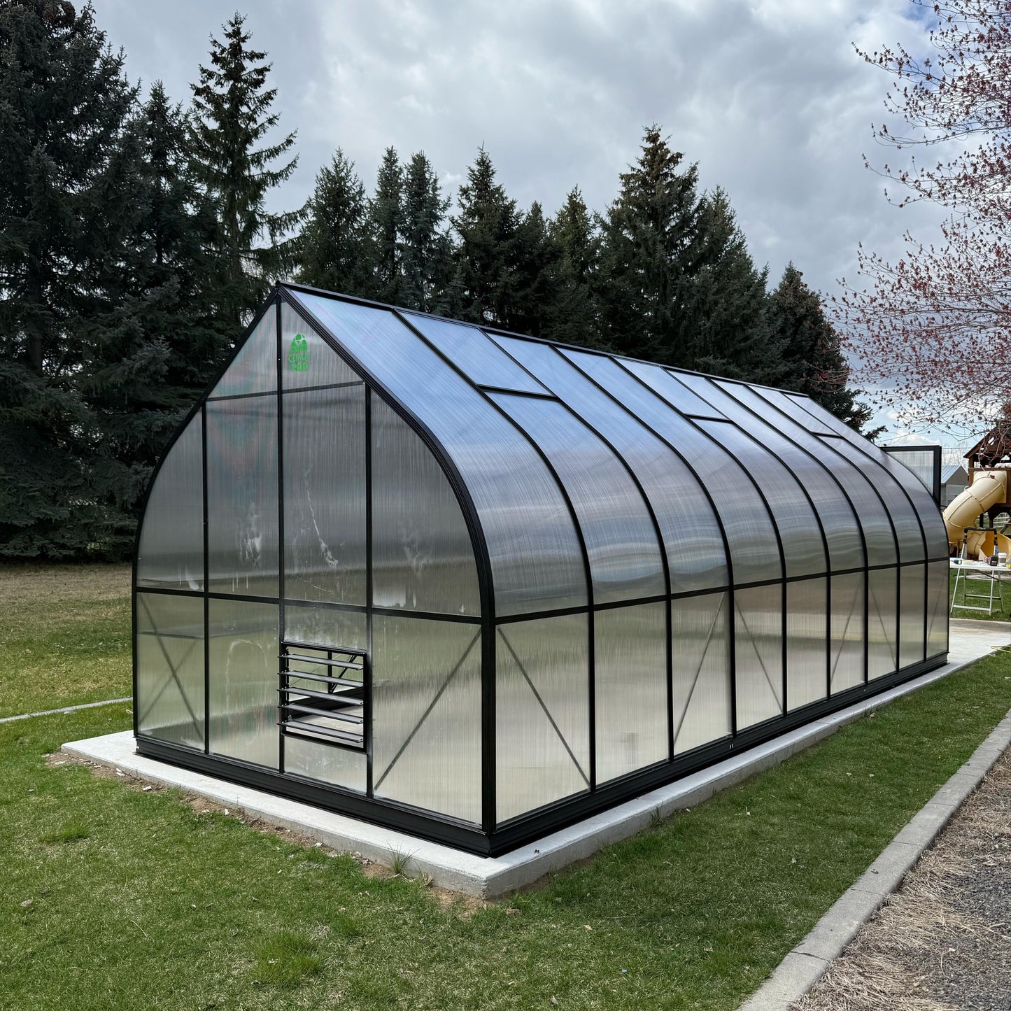 Polycarbonate glass ClimaPod Greenhouse on a grassy area with trees in the background