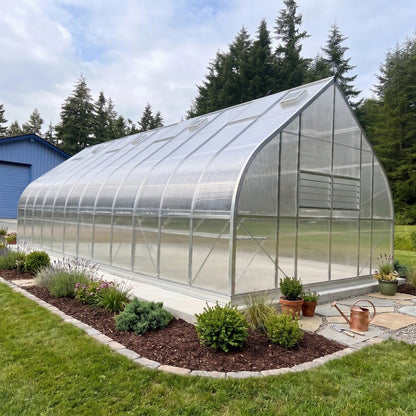 Polycarbonate greenhouse with garden bed in front, surrounded by trees and a blue building